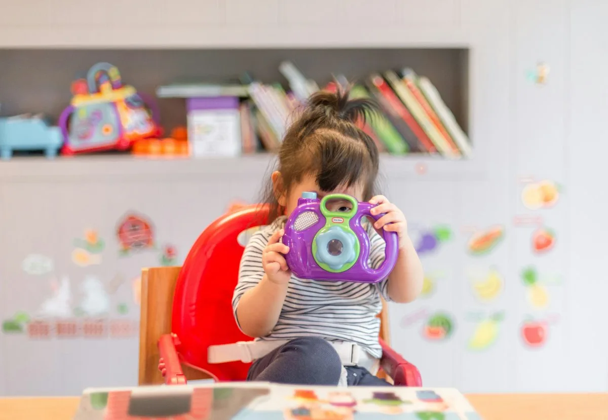 Toddler holding a toy camera in a bright kindergarten room