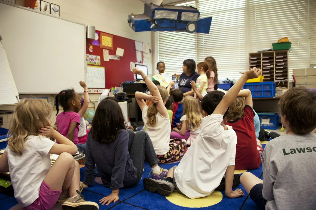 Young children sitting on a classroom carpet raising their hands