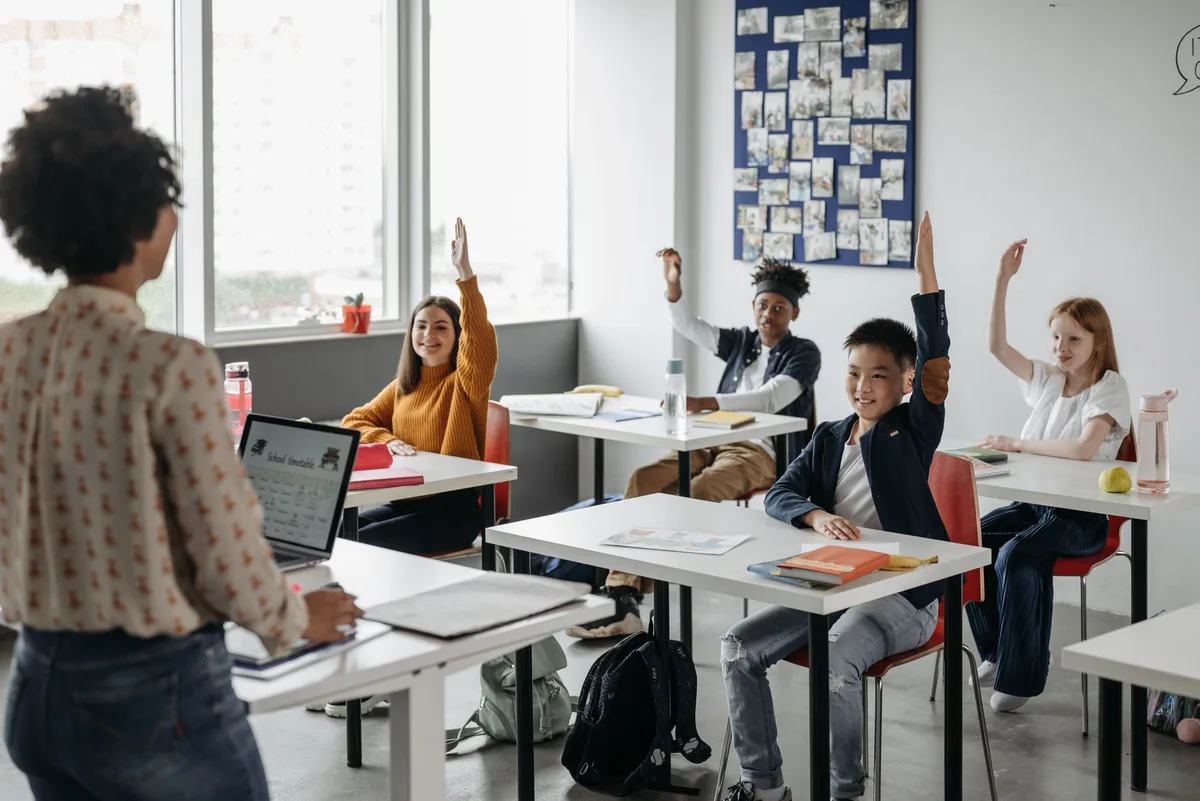 Teacher at the front of a bright classroom while secondary students raise their hands